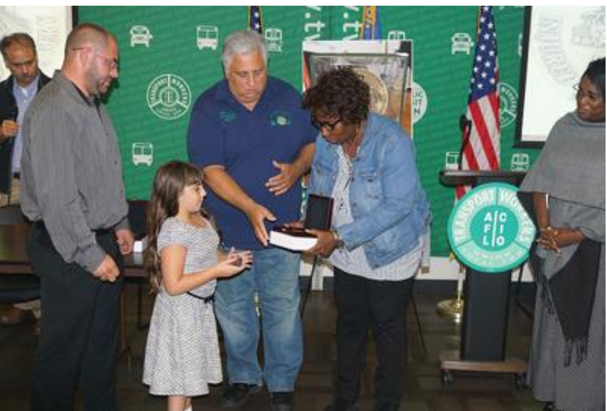 KEEPING FAITH-In 2017 TWU Local 100 recognized members who were part of the 9/11 response-and-recovery efforts at the WTC and now are living with the health consequences of their toxic occupational exposure. Sadie Delman, 8, at the time, is the grand-daughter of a retired TWU Local 100 member James Delman, received the union’s medal given to those with a 9/11 illnesses on his behalf. With her father, at left, are Local 100 President Tony Utano and Shirley Martin.